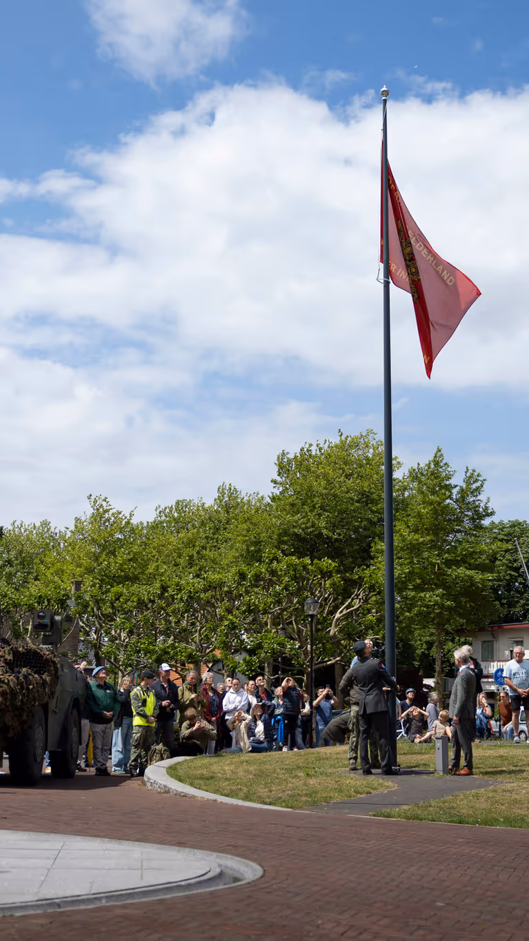Een groep mensen kijkt naar een gecamoufleerde pantserwagen met een rode vlag op een zonnig plein met bomen en huizen.