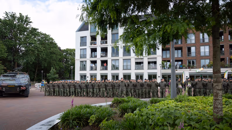 Een rij soldaten in uniform staat in de houding voor een wooncomplex, met een gepantserd voertuig en groen struikgewas.