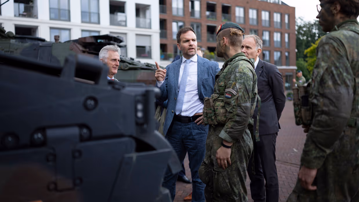 Een man in pak praat met soldaten in camouflage bij een gepantserd voertuig, met woonblokken op de achtergrond.