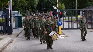Soldaten in camouflage lopen in formatie door een hek, een drummer met trommel vooraan, bomen en gebouwen op de achtergrond. 
