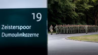 Soldaten in camouflage lopen in formatie langs een weg, met een bord "Zeisterspoor Dumoulinkazerne" en groen bos.
