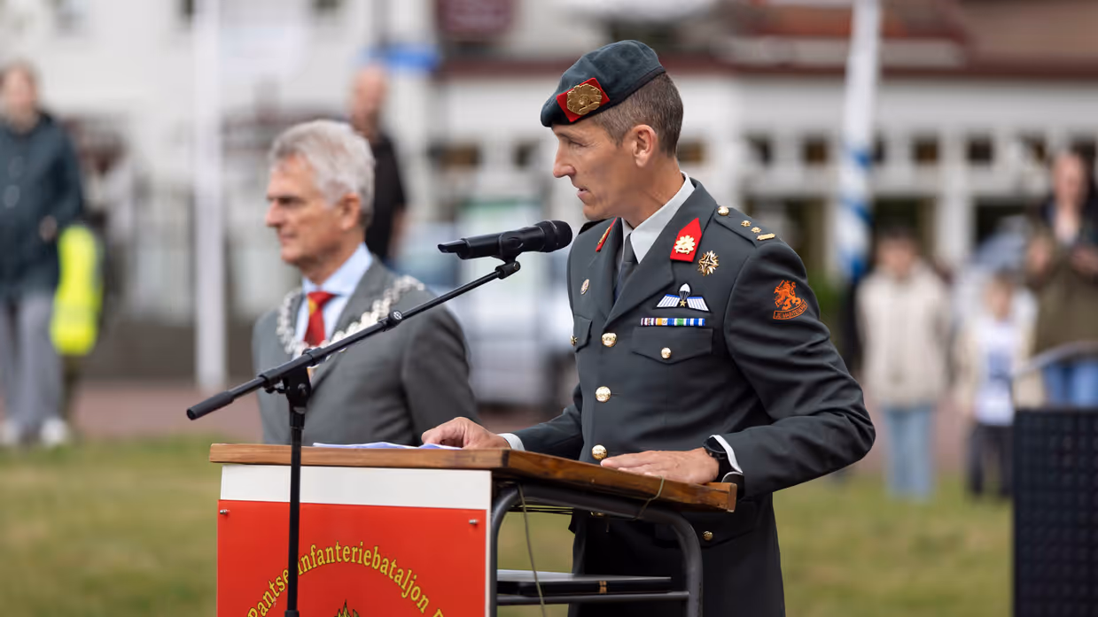 Een man in militair uniform met groene baret houdt een toespraak achter een lessenaar met microfoon, een andere man met ketting kijkt toe.