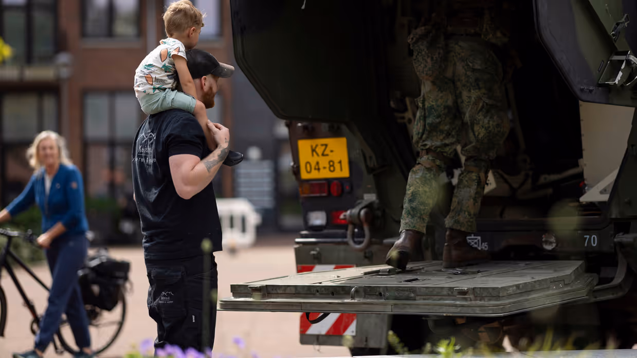 Een man draagt een kind op zijn schouders bij een militaire vrachtwagen, terwijl een soldaat instapt. Op de achtergrond fietst een vrouw.