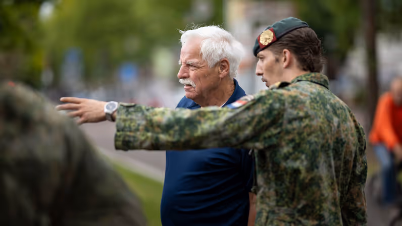 Een oudere man in donkerblauw overhemd en een jongere man in camouflageuniform met groene baret praten buiten, de oudere wijst naar links.