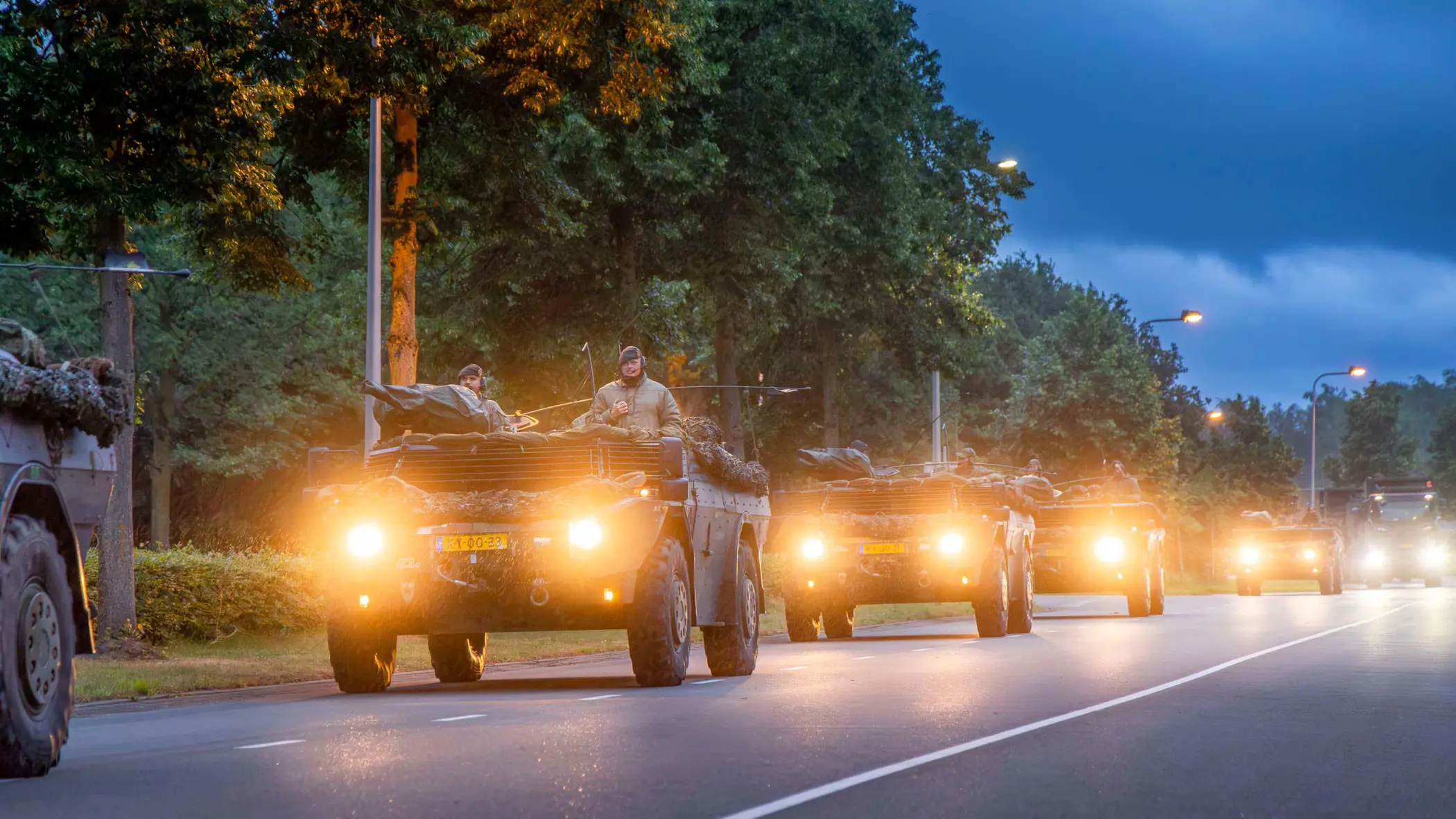 Militaire voertuigen rijden ’s avonds in colonne over een weg.