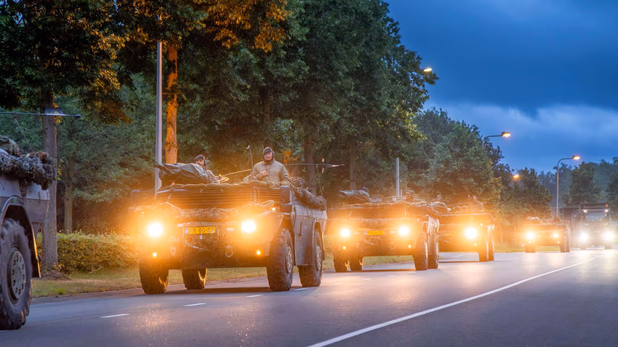 Militaire voertuigen rijden ’s avonds in colonne over een weg.