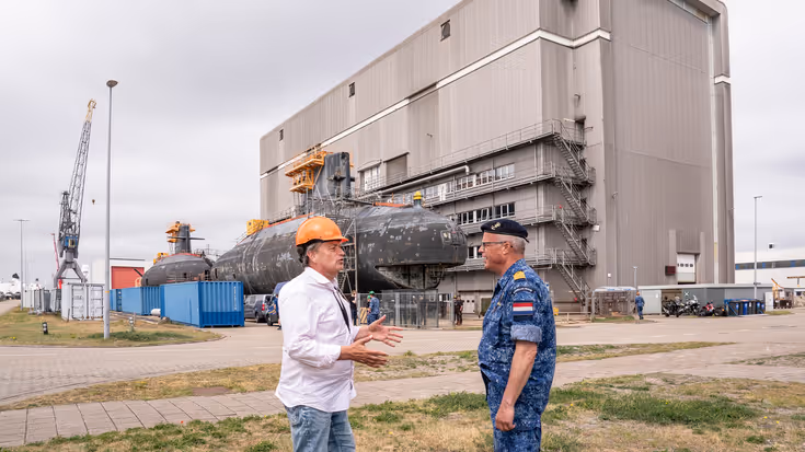 Een burger met oranje bouwhelm op is in gesprek met een marineman, voor een oude onderzeeboot.