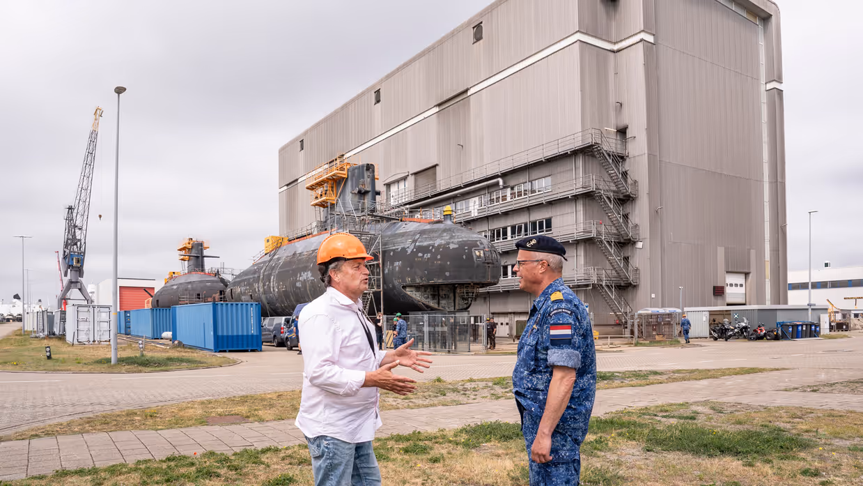 Een burger met oranje bouwhelm op is in gesprek met een marineman, voor een oude onderzeeboot.