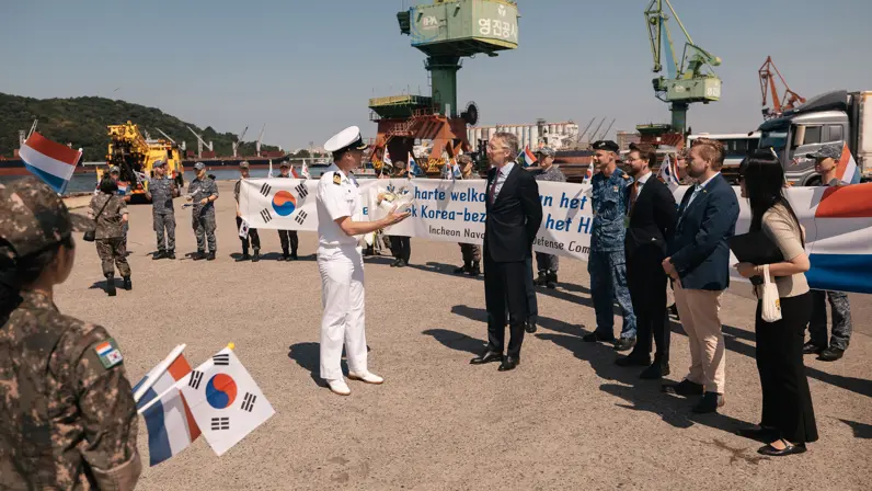 Commandant De Wit schudt handen met iemand van het ontvangstcomité in het Zuid-Koreaanse Incheon. Er wapperen veel Koreaanse vlaggetjes op de kade.