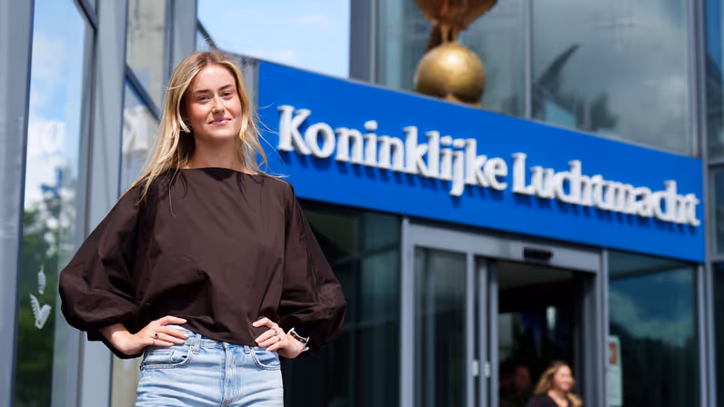  Floor staat voor de ingang van het hoofdkwartier van de Koninklijke Luchtmacht. Op de achtergrond zijn het logo en de ingang van het gebouw met de tekst 'Koninklijke Luchtmacht' zichtbaar.