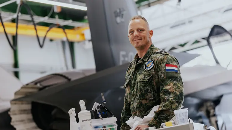 Sergeant 1 Glenn Afman poseert achter een werktafel met kleine spullen erop in een hangaar. Op de achtergrond staat een F-35.
