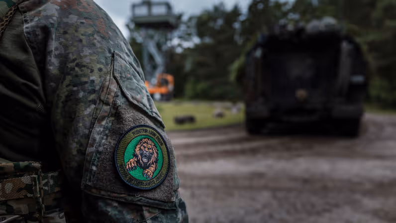 Close-up van de oefenbadge van Fighter Lion 2026 op de mouw van een militair. Op de achtergrond staat een militair voertuig op het oefenterrein.