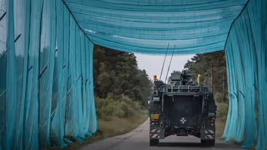 Een militair voertuig rijdt onder een lange strook blauwe camouflagenetten door op een bosweg tijdens oefening Fighter Lion.