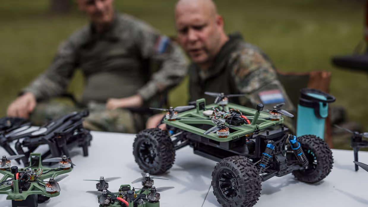 Een onbemand voertuig met een geïntegreerde drone staat op tafel tijdens een demonstratie van TechDevOps, terwijl kapitein Max en luitenant-kolonel Roeland op de achtergrond uitleg geven.
