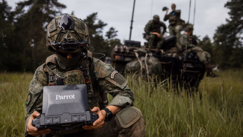 Een militair van 43 Gemechaniseerde Brigade bedient een dronebesturingssysteem terwijl achter hem collega's op een militair voertuig zitten.