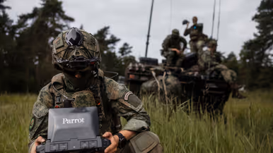Een militair houdt de grondcontroller van een Parrot-drone vast tijdens oefening Fighter Lion, met collega's op een militair voertuig op de achtergrond.