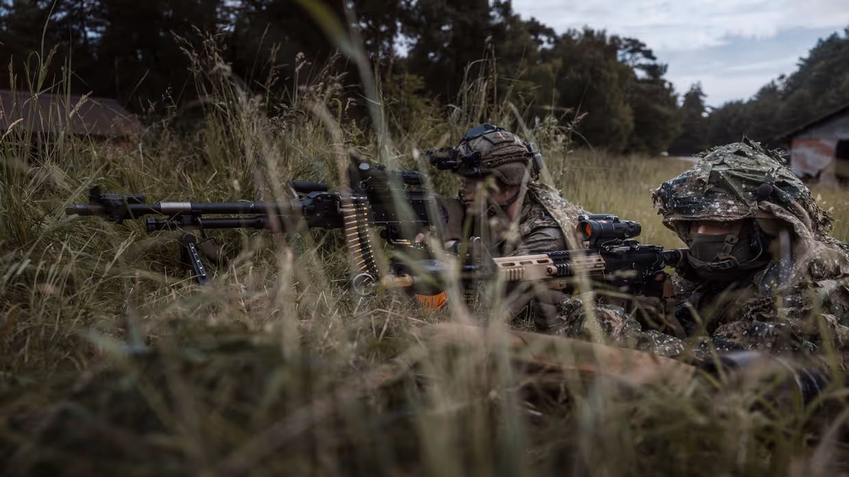 Twee militairen liggen gecamoufleerd in hoog gras tijdens oefening Fighter Lion. Eén bedient een zwaar machinegeweer, terwijl de ander met zijn wapen de omgeving beveiligt. 