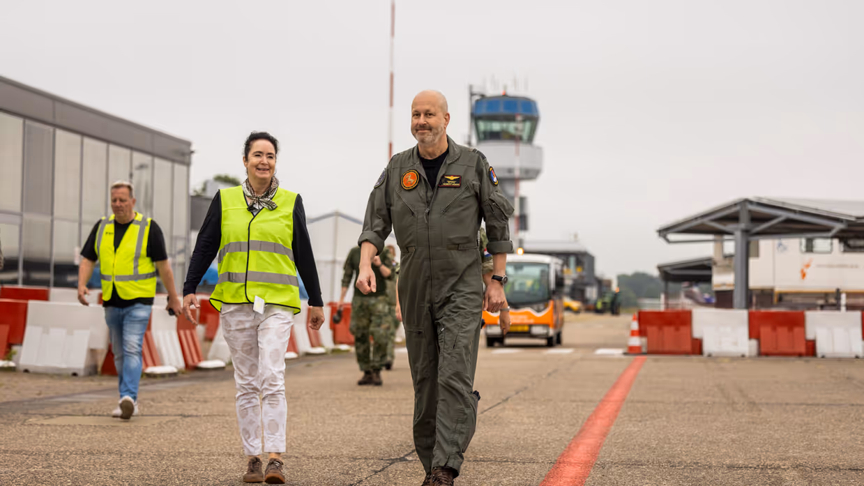 Vrouw in geel hesje loopt naast militair op vliegveld.