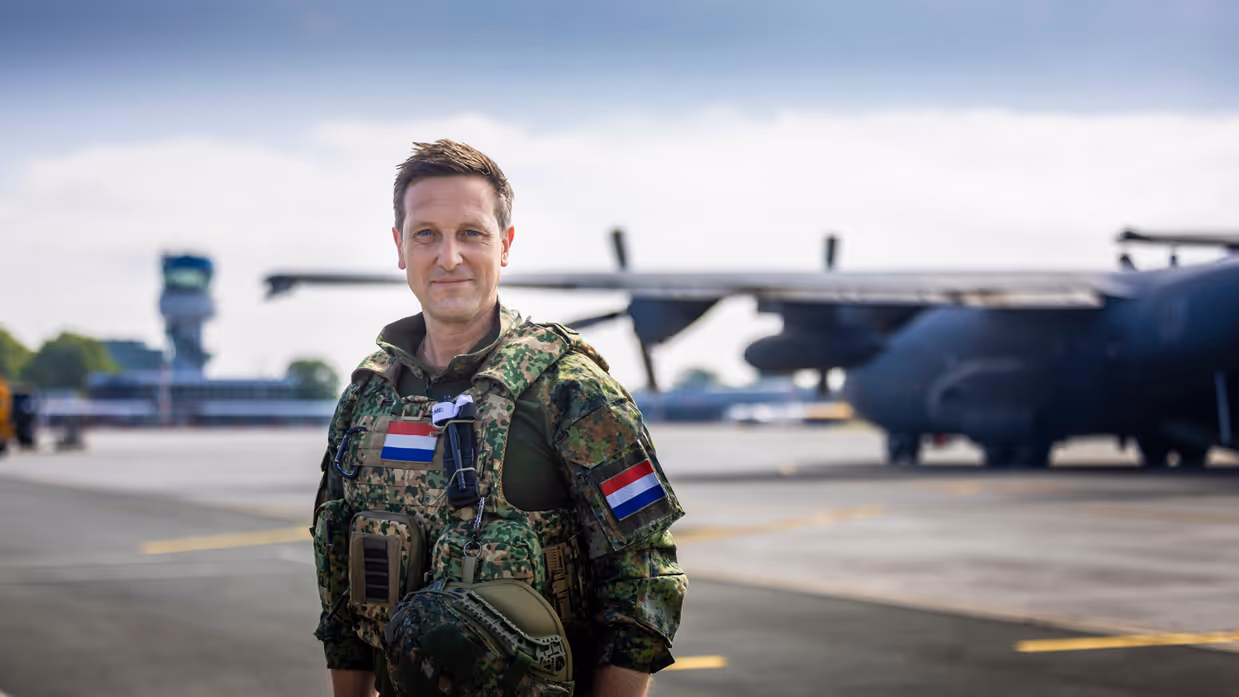 Militair met vest om staat op vliegveld met op de achtergrond een C-130 Hercules-transportvliegtuig.