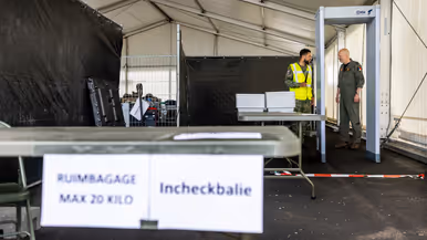 Militair in geel hesje geeft andere militair uitleg bij incheckbalie op tijdelijke vliegbasis.