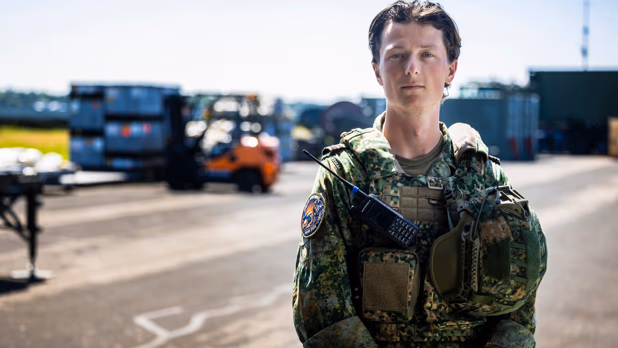 Militair met vest waaraan een helm hangt, staat op taxibaan vliegveld.