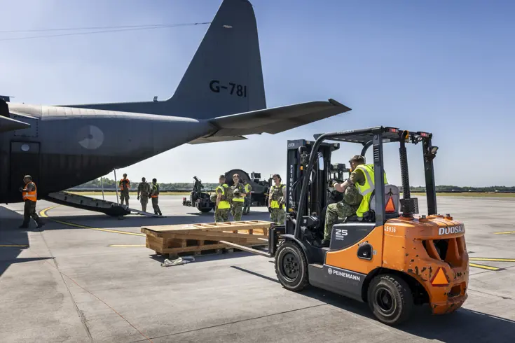 Een vorkheftruck steekt de lepels in een houten stellage. In de achtergrond staat een C-130.