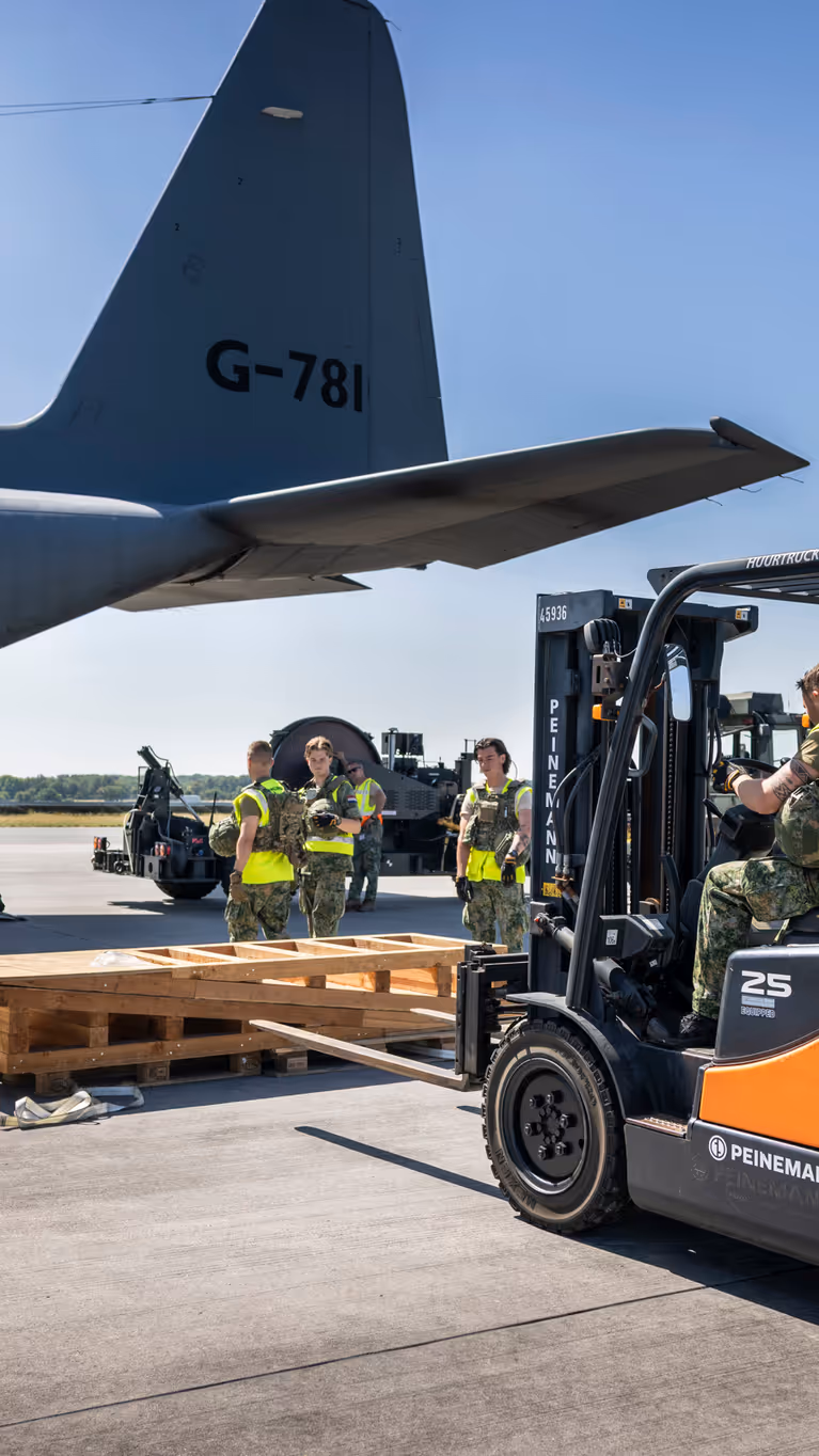 Een vorkheftruck steekt de lepels in een houten stellage. In de achtergrond staat een C-130.