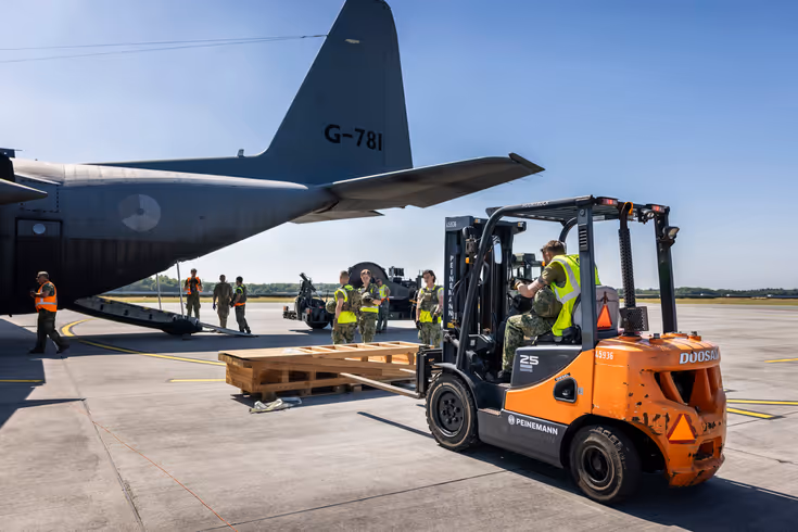 Een vorkheftruck steekt de lepels in een houten stellage. In de achtergrond staat een C-130.