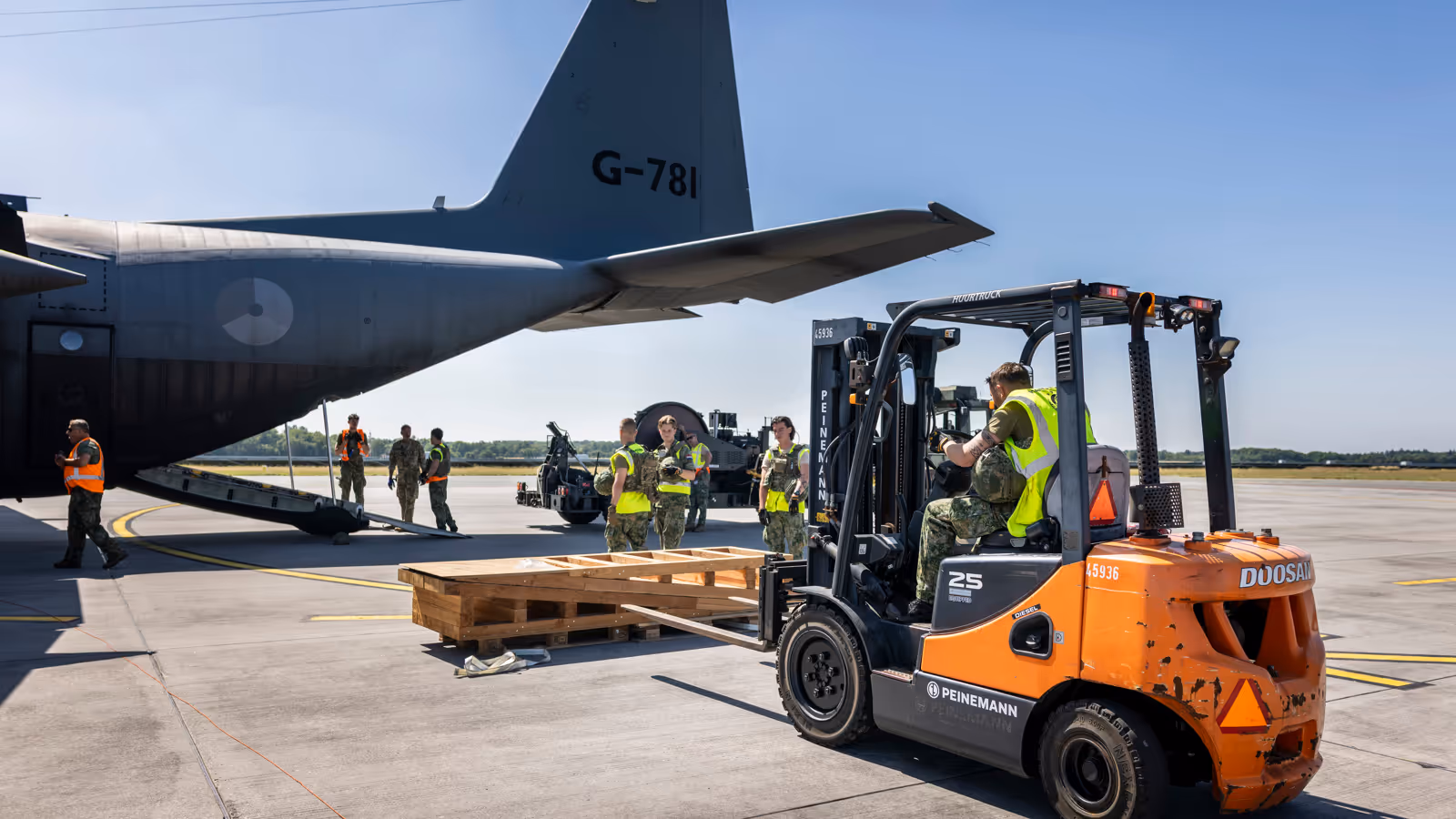 Een vorkheftruck steekt de lepels in een houten stellage. In de achtergrond staat een C-130.
