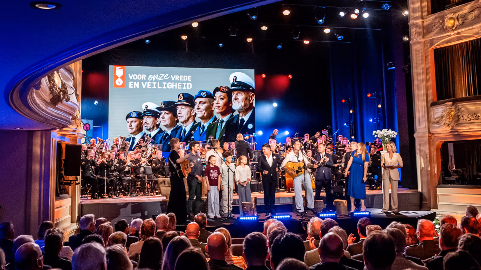 De finale van de ochtend met alle solisten, sprekers, presentatrice en een aantal kinderen op het podium. In de achtergrond is boven het orkest groot een afbeelding van Nederlandse veteranen te zien. 