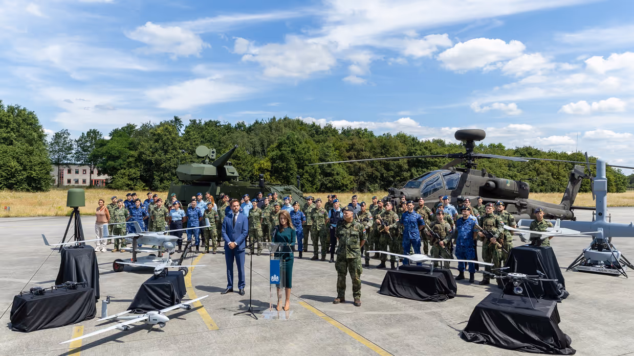 De minister presenteert de Defensienota met naast haar de stattssecretaris en de Commandant der Strijdkrachten. Achter hen staat een brede delegatie militairen en een Apache-gevechtshelikopter.