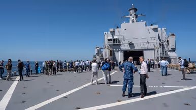 het achterdek van een marineschip staat vol met mensen die met elkaar praten.