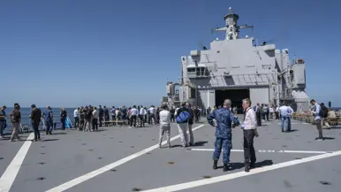 het achterdek van een marineschip staat vol met mensen die met elkaar praten.