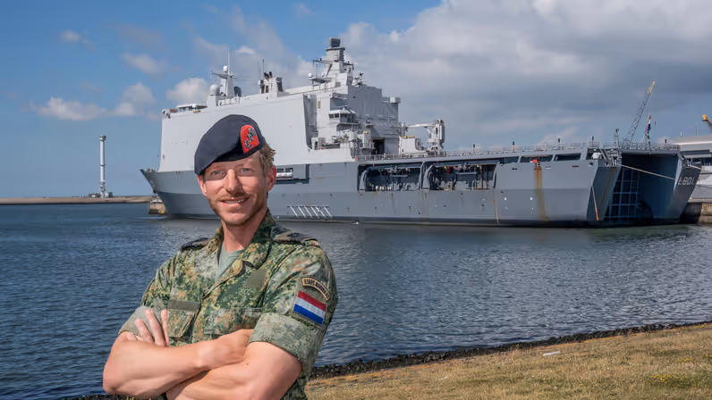 Marinier met baret op de kade met handen over elkaar. Op de achtergrond een groot marineschip.