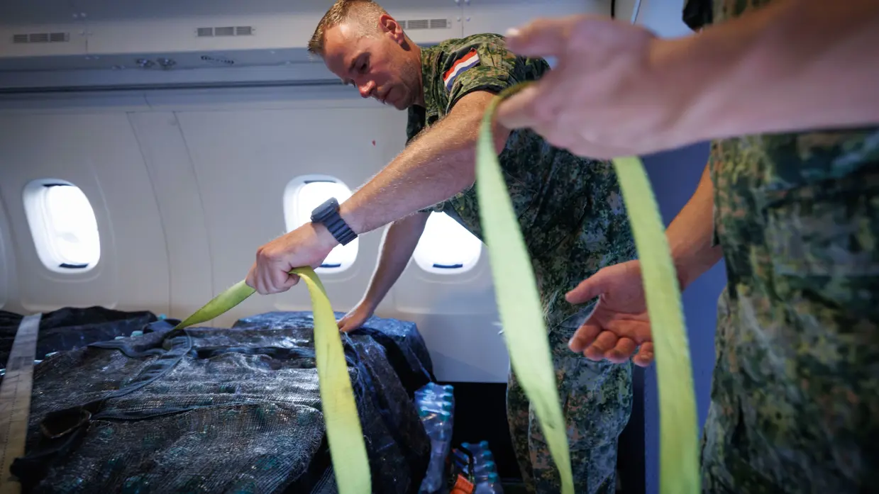 Een militair staat in het laadruim van een vliegtuig. Hij sjort aan een gele spanband om een vracht vast te zetten.