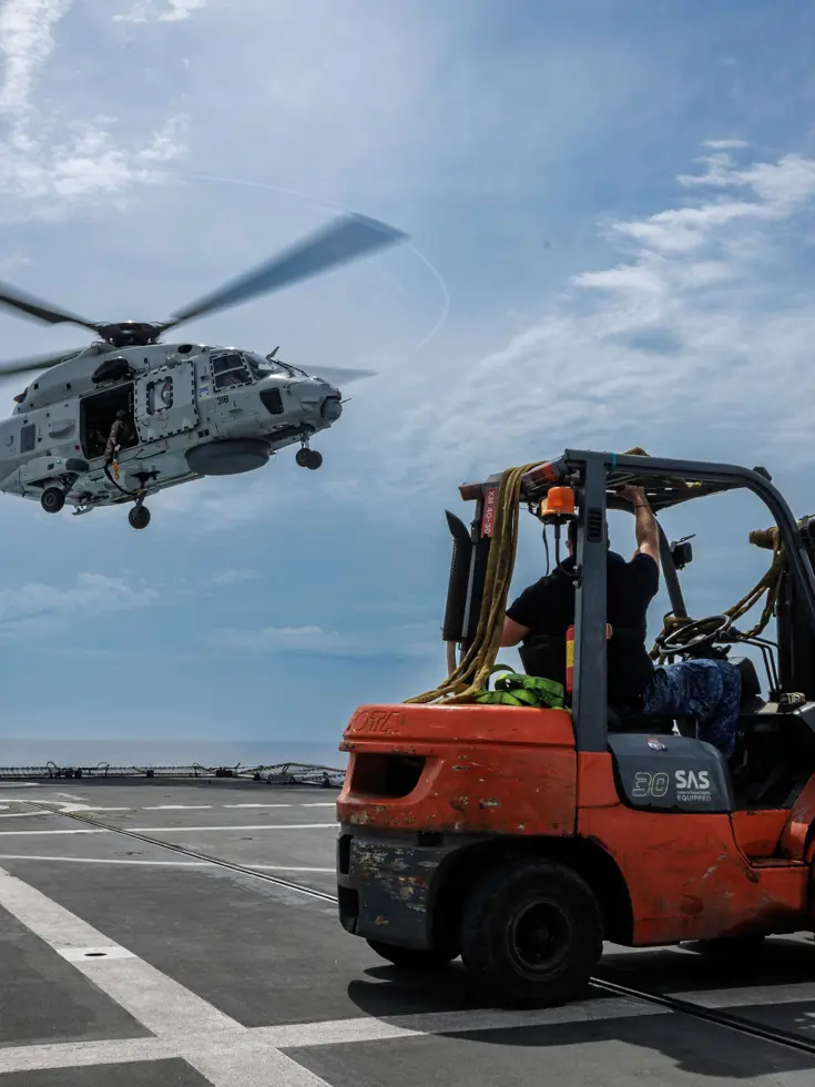 Een NH90 hangt boven het helidek. Links staat een pallet hulpgoederen, rechts staat een vorkheftruck met een volgende lading.