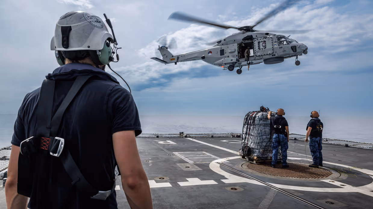 Een marinecollega kijkt naar een helikopter die boven een scheepsdek hangt.