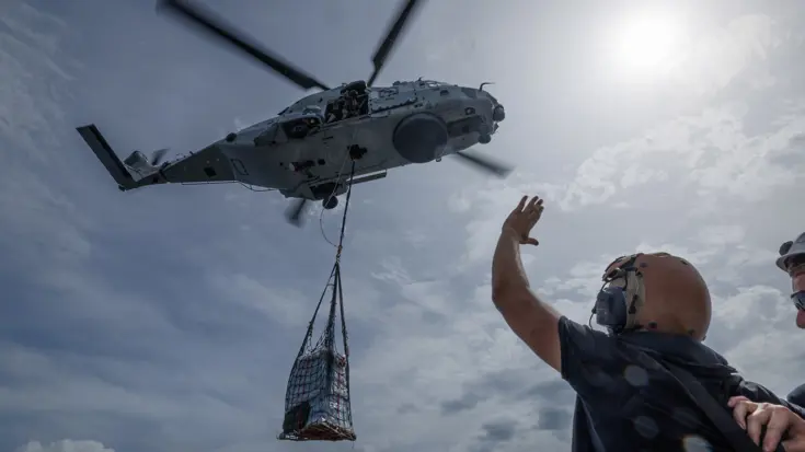 Een helikopter vliegt weg met een lading hulpgoederen hangend onder het toestel. Vanaf het helidek zwaait een militair naar de helikopter.