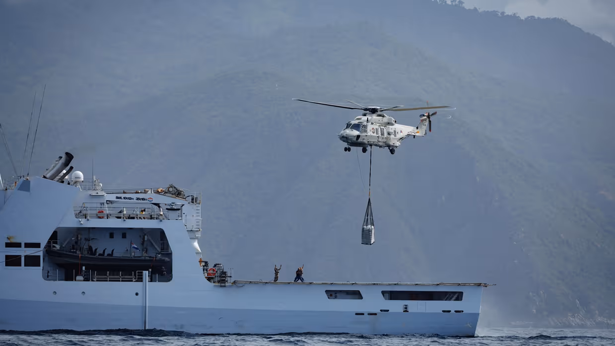 Er hangt een helikopter boven het achterdek en tilt een vracht de lucht in. 