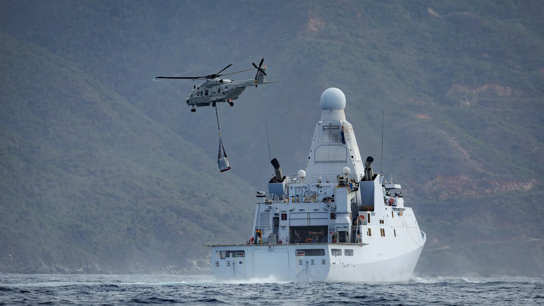 Een NH90 vliegt met een lading aan een net onder de helikopter weg van een marineschip