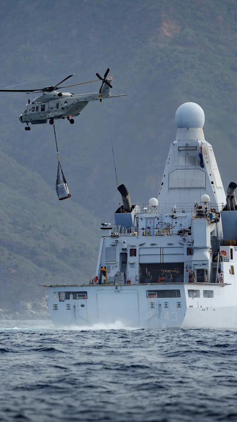 Een NH90 vliegt met een lading aan een net onder de helikopter weg van een marineschip
