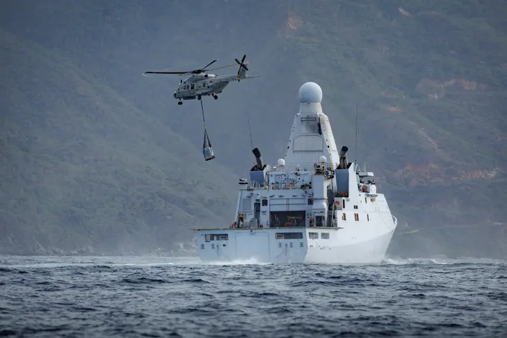 Een NH90 vliegt met een lading aan een net onder de helikopter weg van een marineschip