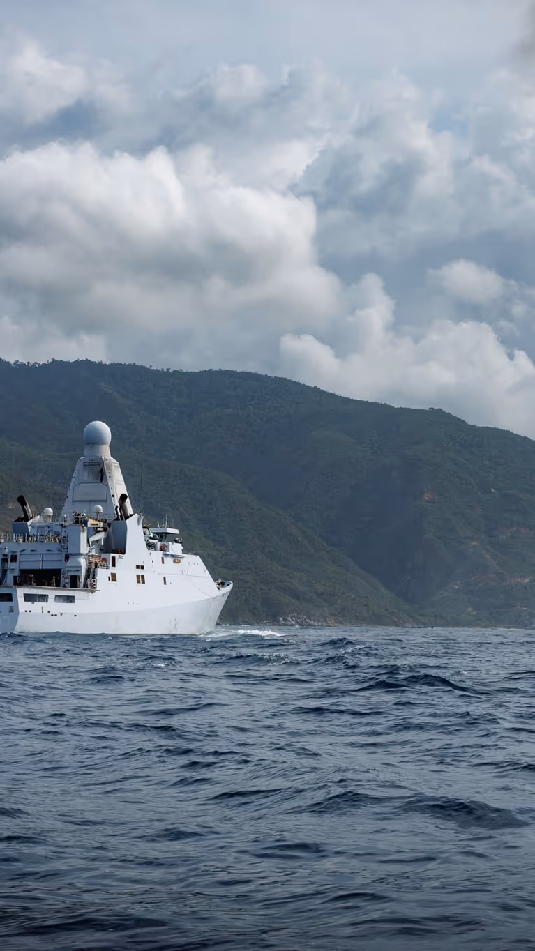 Een marinecollega kijkt hoe in de verte een helikopter een vracht oppakt van een marineschip.