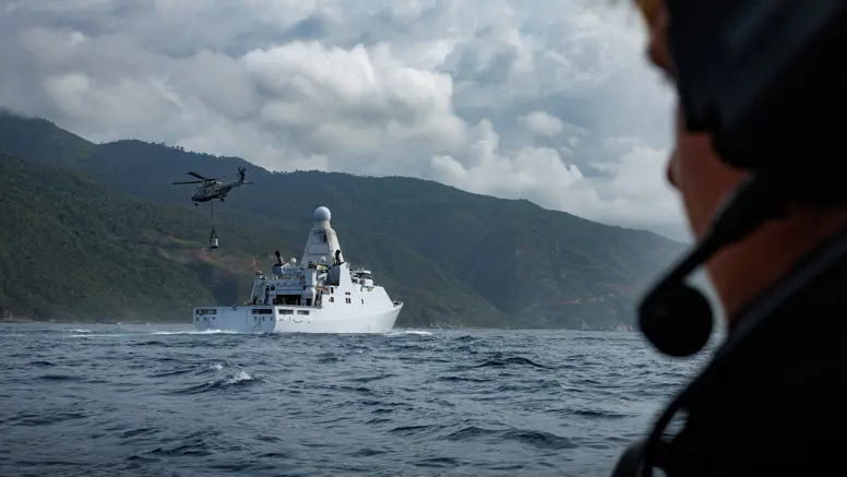 Een marinecollega kijkt hoe in de verte een helikopter een vracht oppakt van een marineschip.