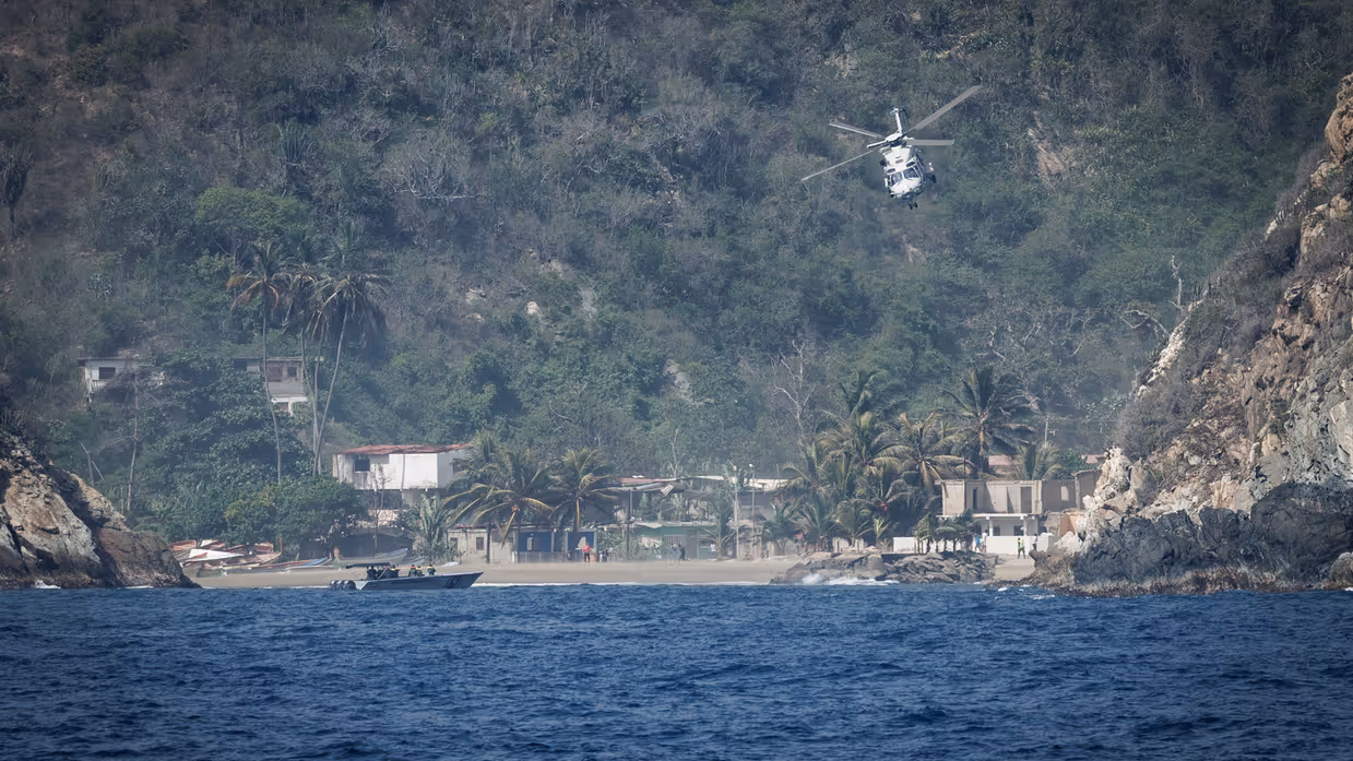 Een helikopter vliegt richting de lens, op de achtergrond groene bergen en wat huisjes.