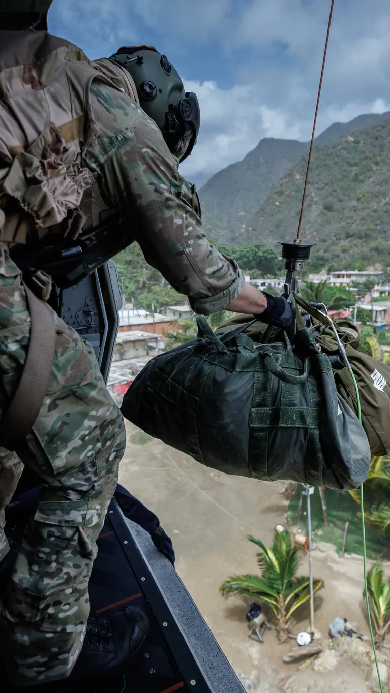 Een bemanningslid van de NH90 staat in de deuropening en is bezig met een vrachtbundel die aan een kabel buiten het toestel hangt.