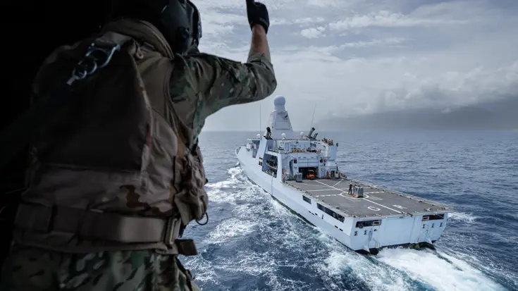 Een bemanningslid van een NH90 steekt zijn duim op terwijl de helikopter langs Zr.Ms. Groningen vliegt.