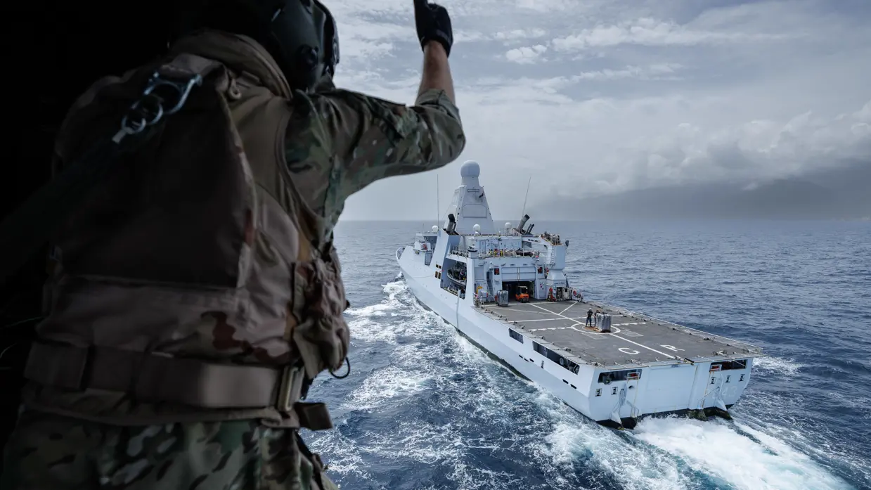 Een bemanningslid van een NH90 steekt zijn duim op terwijl de helikopter langs Zr.Ms. Groningen vliegt.