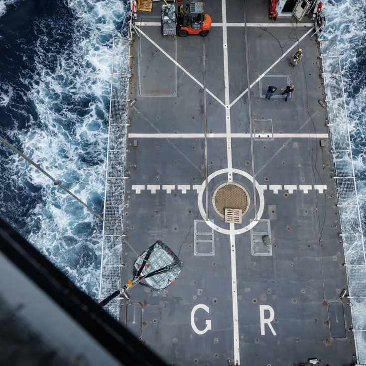 Het grijze helikdek gezien vanuit de helikopter, terwijl het schip vaart.