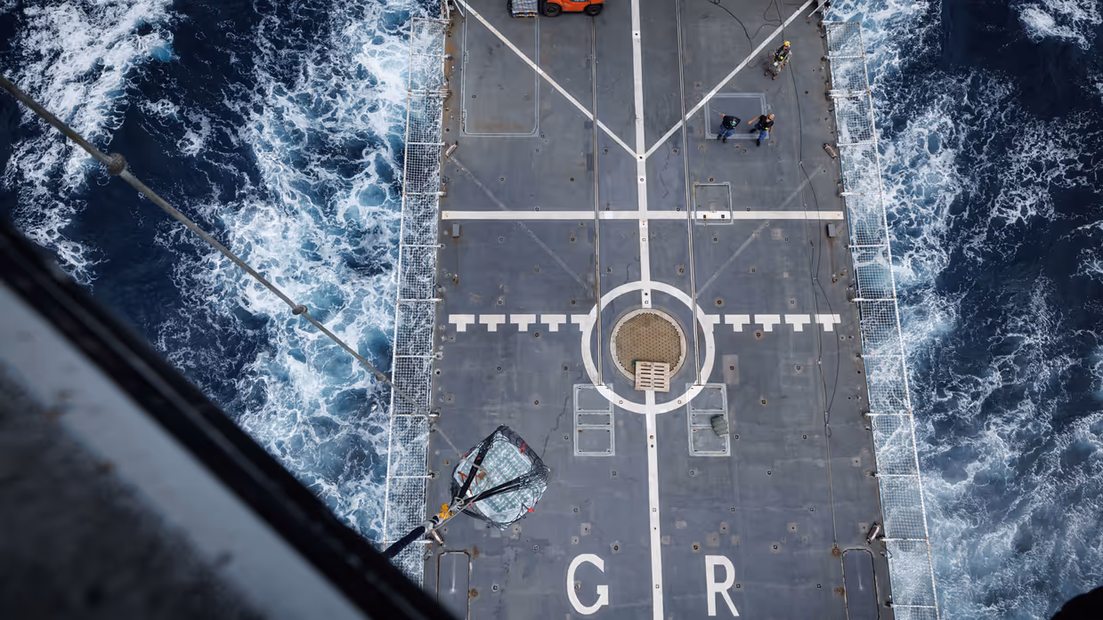 Het grijze helikdek gezien vanuit de helikopter, terwijl het schip vaart.